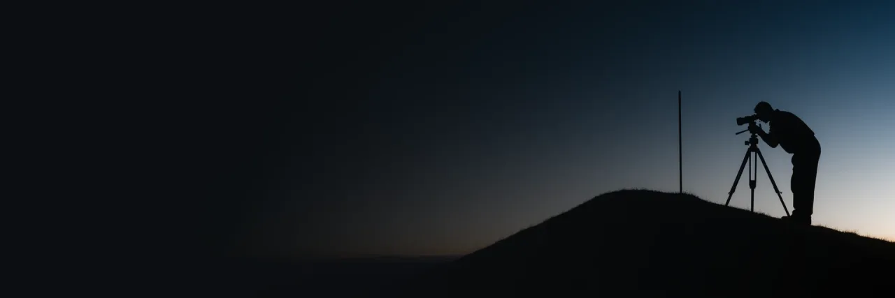 A land surveyor in silhouette bent to the eyepiece of a theodolite on a tripod atop a low hill, a ranging pole standing nearby, against a deep blue dusk sky